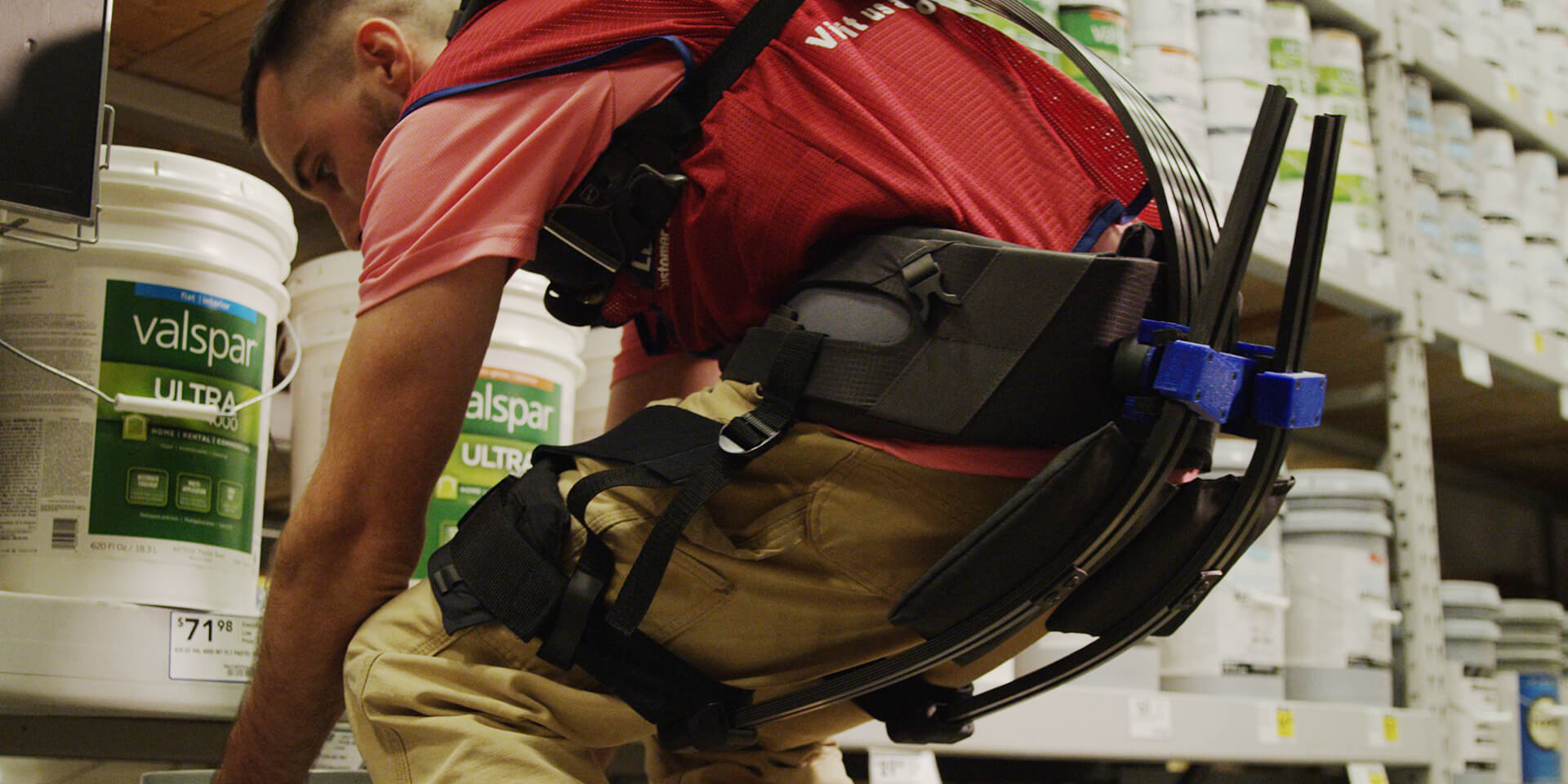 Lowe's Home Improvement associate on sales floor lifting heavy object while wearing soft robotic exoskeleton for assistance.