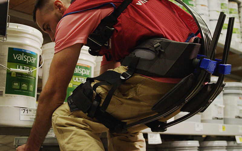 Lowe's Home Improvement associate on sales floor lifting heavy object while wearing soft robotic exoskeleton for assistance.